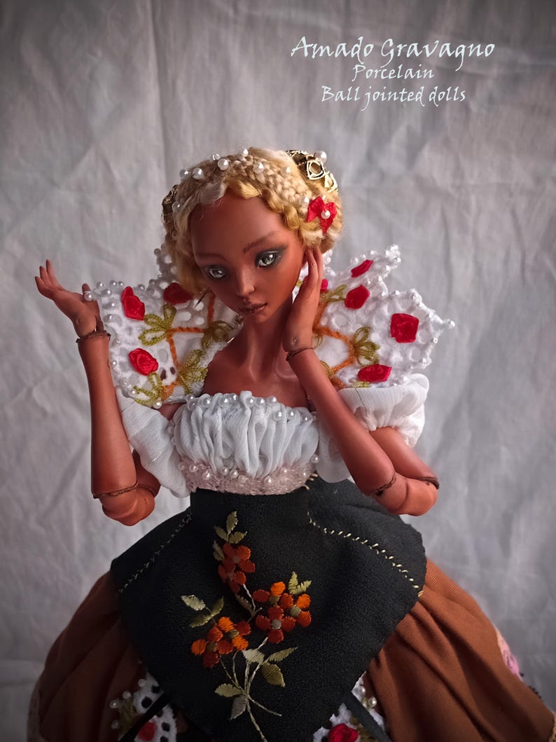 Pode incluir: Uma boneca de porcelana articulada com cabelo loiro tran&ccedil;ado adornado com p&eacute;rolas e la&ccedil;os vermelhos. A boneca usa um top branco com babados e p&eacute;rolas, um grande colarinho de renda branca com detalhes florais vermelhos e um avental preto sobre saia castanha.