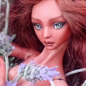 Pode incluir: Uma boneca articulada de porcelana com longos cabelos castanho-avermelhados e olhos verde-claros, rodeada por ramos de lavanda roxa. O texto 'Amado Gravagno Porcelain Ball jointed dolls' &eacute; vis&iacute;vel no canto superior direito da imagem.