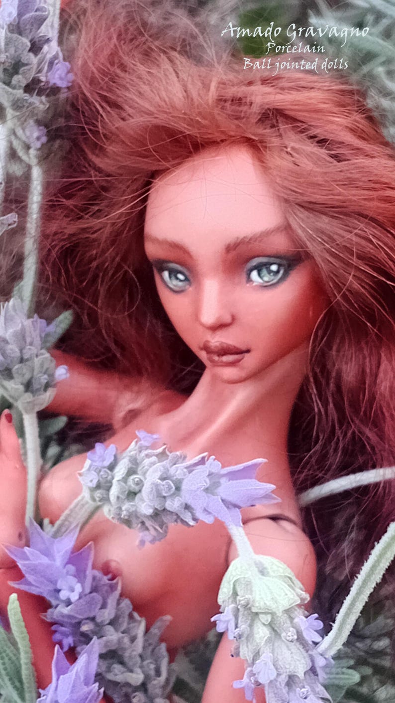 Pode incluir: Uma boneca articulada de porcelana com longos cabelos castanho-avermelhados e olhos verde-claros, rodeada por ramos de lavanda roxa. O texto 'Amado Gravagno Porcelain Ball jointed dolls' &eacute; vis&iacute;vel no canto superior direito da imagem.
