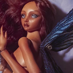 Pode incluir: Uma boneca articulada de porcelana com pele bronzeada, cabelos longos e ondulados escuros e olhos azul-claros. A boneca apresenta asas texturizadas azul-escuras e est&aacute; marcada com o texto Amado Gravagno Porcelain Ball jointed dolls no canto.