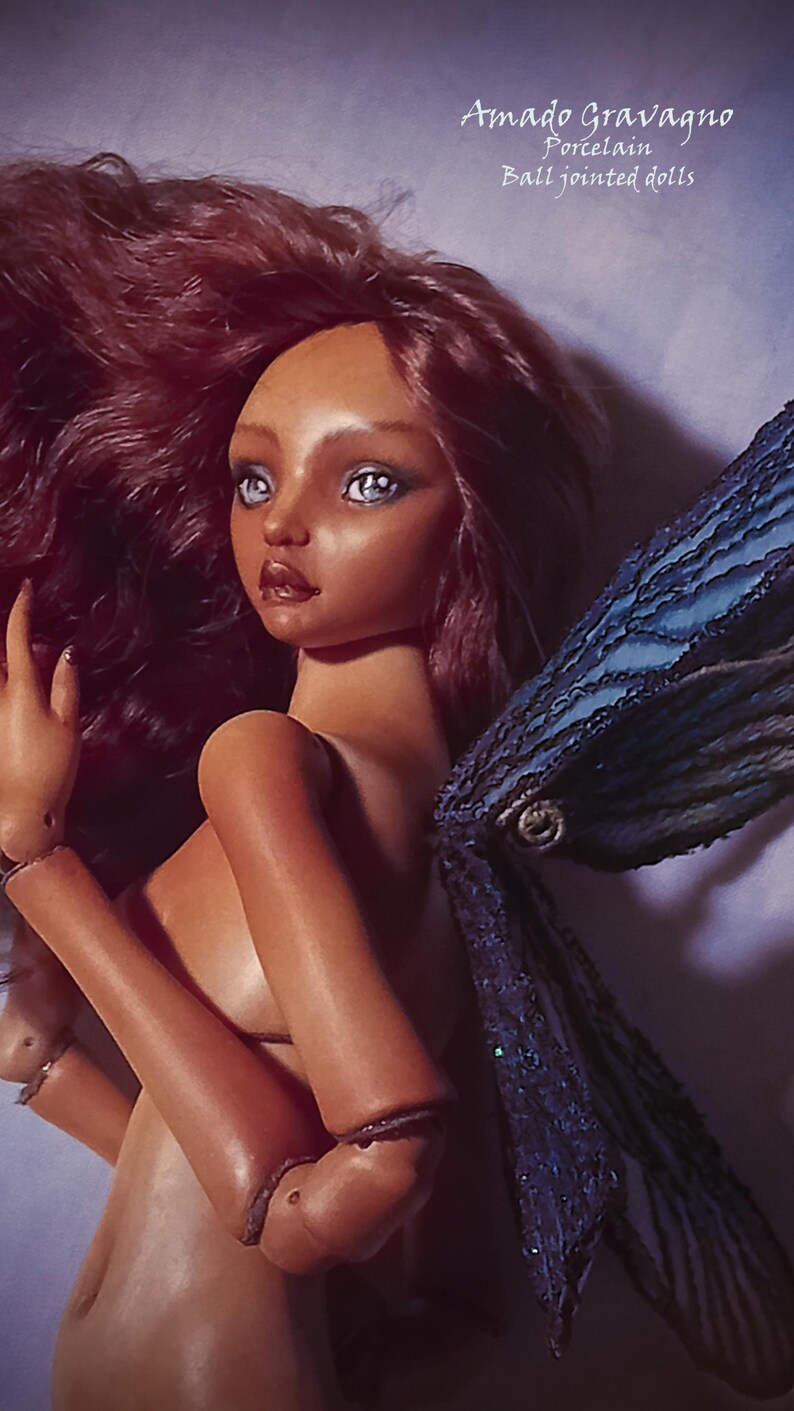 Pode incluir: Uma boneca articulada de porcelana com pele bronzeada, cabelos longos e ondulados escuros e olhos azul-claros. A boneca apresenta asas texturizadas azul-escuras e est&aacute; marcada com o texto Amado Gravagno Porcelain Ball jointed dolls no canto.