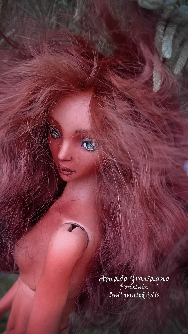 Pode incluir: Uma boneca articulada de pele bronzeada, com longos cabelos ondulados castanho-avermelhados e olhos verde-claros. A boneca apresenta uma articula&ccedil;&atilde;o de ombro vis&iacute;vel e o texto Amado Gravagno Porcelain Ball jointed dolls no canto inferior.