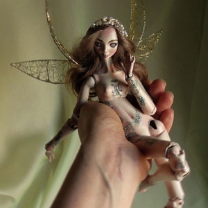 May include: A porcelain doll with wings and tattoos, wearing a crown, is held in a hand. The doll has long brown hair and blue eyes. The doll is a handmade art piece by Amado-Gravagno Porcelain Dolls.