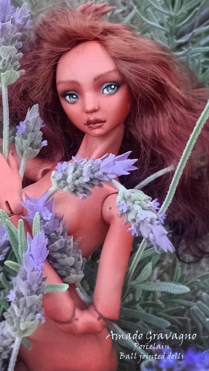 Pode incluir: Uma boneca articulada de pele bronzeada, com longos cabelos ondulados castanho-avermelhados e olhos azul-claros, posicionada entre ramos de lavanda roxa. O texto "Amado Gravagno Porcelain Ball jointed dolls" est&aacute; impresso em branco no canto inferior direito.