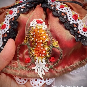Pode incluir: Uma pe&ccedil;a de cetim cor de ferrugem com bordados intrincados de motivos florais laranja, missangas brancas e detalhes em fio verde. A pe&ccedil;a &eacute; adornada com renda preta e branca, pequenas rosas de fita vermelha e uma borda de renda recortada.