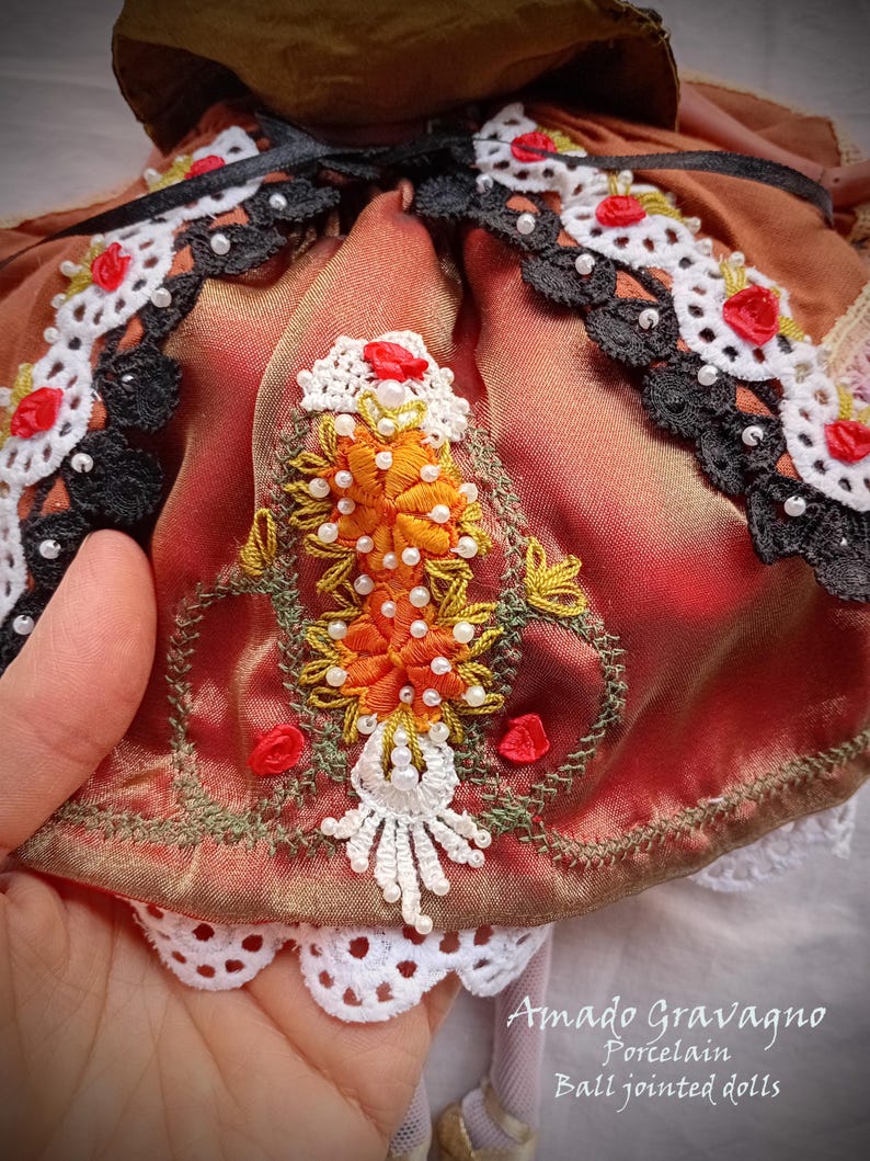 Pode incluir: Uma pe&ccedil;a de cetim cor de ferrugem com bordados intrincados de motivos florais laranja, missangas brancas e detalhes em fio verde. A pe&ccedil;a &eacute; adornada com renda preta e branca, pequenas rosas de fita vermelha e uma borda de renda recortada.