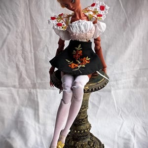 Pode incluir: Uma boneca articulada de pele escura e cabelo loiro adornado com p&eacute;rolas. A boneca veste um top branco de ombros ca&iacute;dos com renda e detalhes florais vermelhos, uma saia preta com bordado floral laranja, meias arrast&atilde;o brancas e sapatos amarelos.