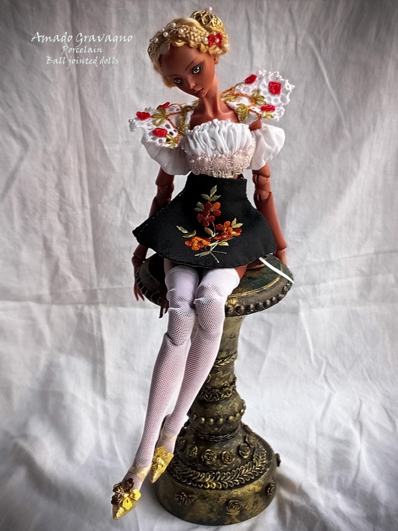 Pode incluir: Uma boneca articulada de pele escura e cabelo loiro adornado com p&eacute;rolas. A boneca veste um top branco de ombros ca&iacute;dos com renda e detalhes florais vermelhos, uma saia preta com bordado floral laranja, meias arrast&atilde;o brancas e sapatos amarelos.