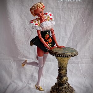Pode incluir: Uma boneca de porcelana articulada com pele bronzeada, cabelo loiro e um conjunto preto e branco com bordados florais. A boneca usa meias arrast&atilde;o brancas e sapatos dourados, em p&eacute; ao lado de um pedestal dourado de estilo antigo.