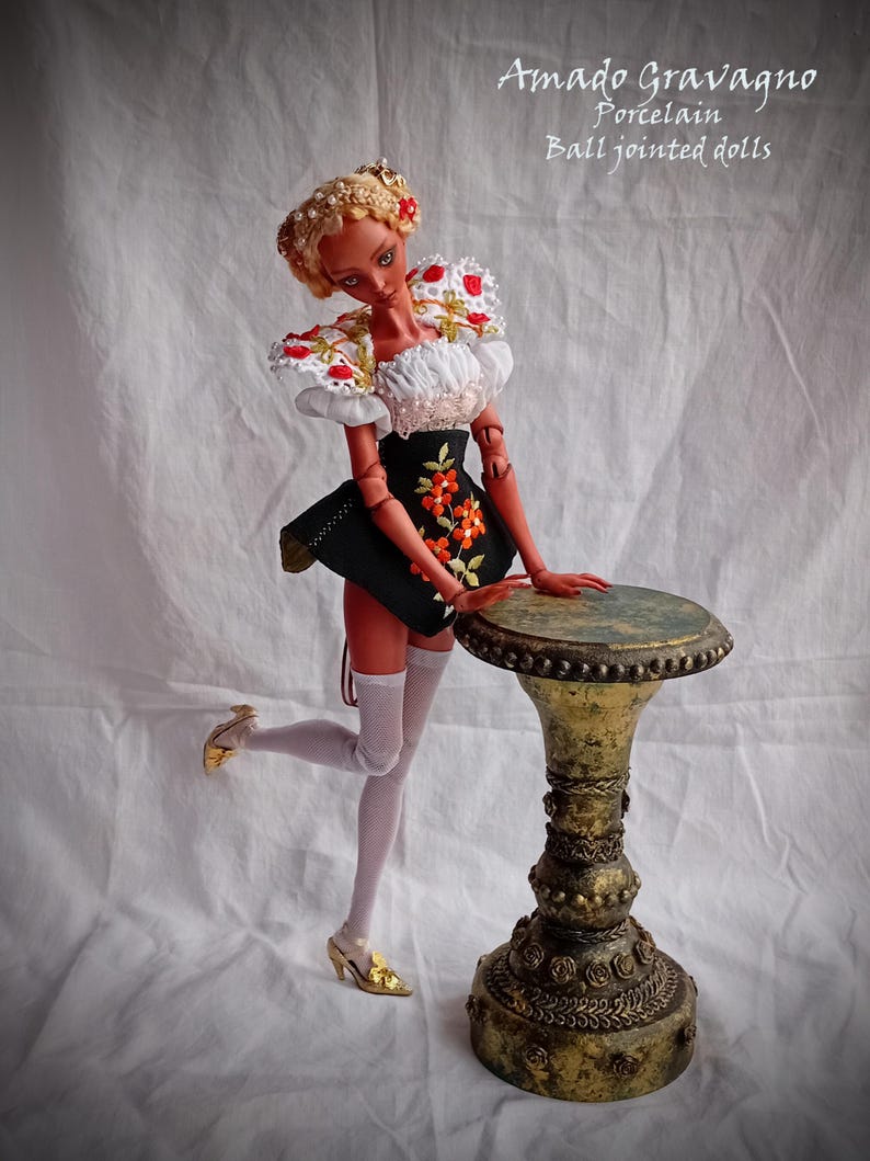 Pode incluir: Uma boneca de porcelana articulada com pele bronzeada, cabelo loiro e um conjunto preto e branco com bordados florais. A boneca usa meias arrast&atilde;o brancas e sapatos dourados, em p&eacute; ao lado de um pedestal dourado de estilo antigo.