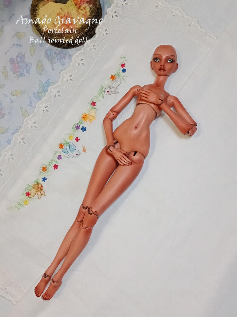 Pode incluir: Uma boneca articulada de porcelana com pele bronzeada, membros m&oacute;veis, olhos escuros e acabamento liso. A boneca est&aacute; deitada, com articula&ccedil;&otilde;es vis&iacute;veis nos ombros, cotovelos, pulsos, quadris, joelhos e tornozelos. O texto diz Amado Gravagno Porcelain Ball jointed dolls.