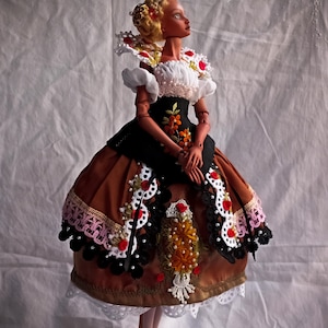 Pode incluir: Uma boneca articulada de porcelana com cabelo loiro, vestindo um traje castanho e preto com renda branca, detalhes florais vermelhos e bordados. O texto Amado Gravagno Porcelain Ball jointed dolls &eacute; vis&iacute;vel no canto superior direito.