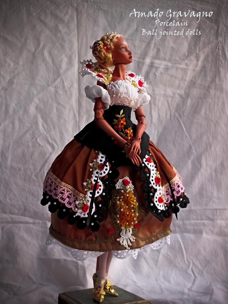 Pode incluir: Uma boneca articulada de porcelana com cabelo loiro, vestindo um traje castanho e preto com renda branca, detalhes florais vermelhos e bordados. O texto Amado Gravagno Porcelain Ball jointed dolls &eacute; vis&iacute;vel no canto superior direito.