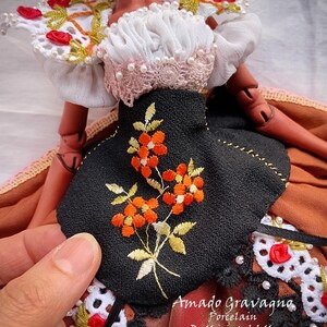 Pode incluir: Uma boneca articulada vestindo uma blusa branca com detalhes em renda e p&eacute;rolas, um corpete preto com bordado floral laranja e uma saia marrom. O texto Amado Gravagno Porcelain Ball jointed dolls &eacute; vis&iacute;vel no canto inferior direito.