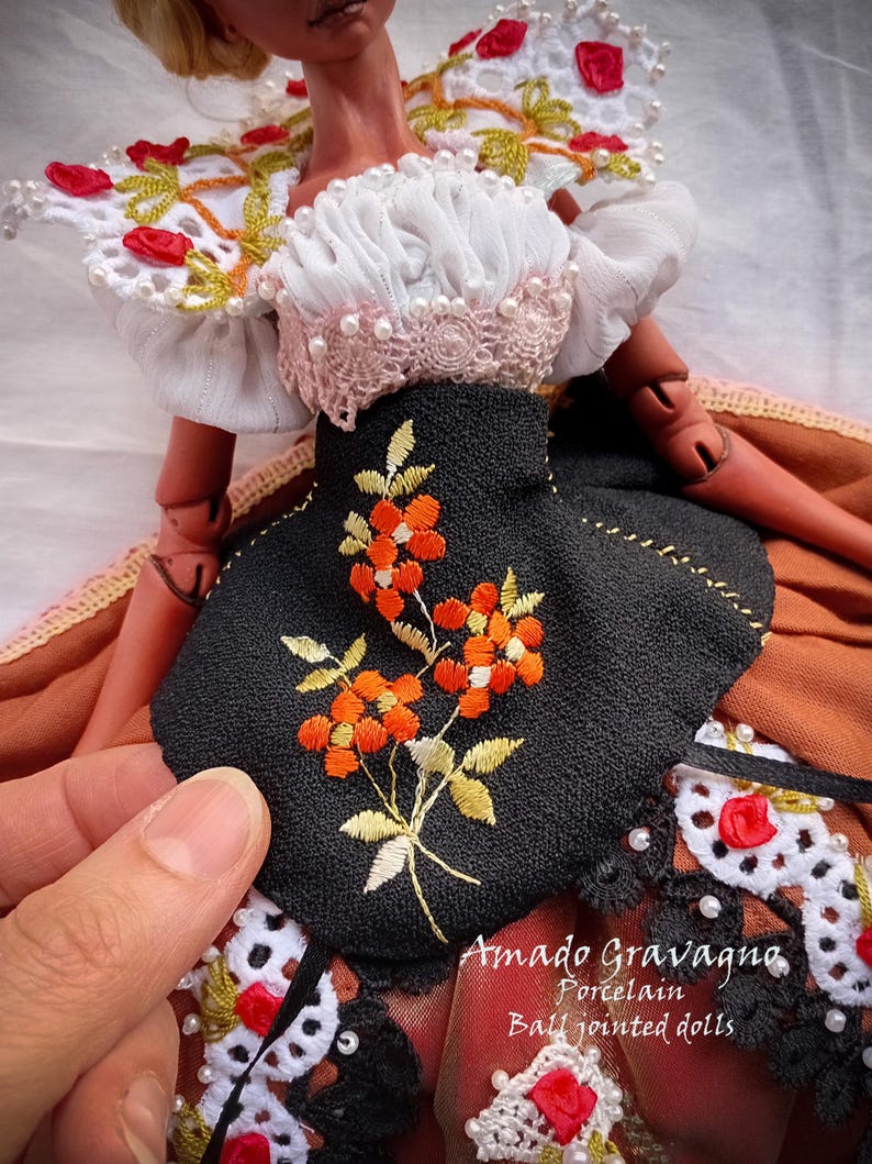Pode incluir: Uma boneca articulada vestindo uma blusa branca com detalhes em renda e p&eacute;rolas, um corpete preto com bordado floral laranja e uma saia marrom. O texto Amado Gravagno Porcelain Ball jointed dolls &eacute; vis&iacute;vel no canto inferior direito.