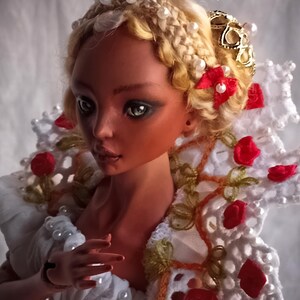 Pode incluir: Uma boneca articulada de porcelana com pele bronzeada e cabelo loiro em uma coroa tran&ccedil;ada com contas brancas. A boneca usa um colarinho de renda branca adornado com pequenas rosas de tecido vermelho e p&eacute;rolas, com o texto Amado Gravagno Porcelain Ball jointed dolls.