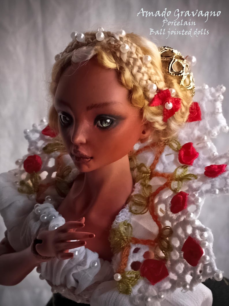 Pode incluir: Uma boneca articulada de porcelana com pele bronzeada e cabelo loiro em uma coroa tran&ccedil;ada com contas brancas. A boneca usa um colarinho de renda branca adornado com pequenas rosas de tecido vermelho e p&eacute;rolas, com o texto Amado Gravagno Porcelain Ball jointed dolls.