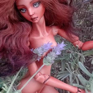Pode incluir: Uma boneca articulada com pele castanho-avermelhada e longos cabelos castanhos ondulados, com olhos claros e membros articulados. A boneca segura um ramo de lavanda roxa. O texto na parte inferior diz Amado Gravagno Porcelain Ball jointed dolls.