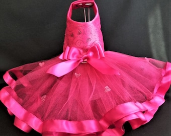May include: A pink tulle tutu dress for a pet with a pink satin bow and a pink satin trim. The dress has a sparkly pink bodice with a swirl design.