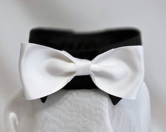 May include: A formal black collar with a white satin bow tie. The bow tie is centered on the collar and is tied in a classic bow shape. The collar has two small black points.