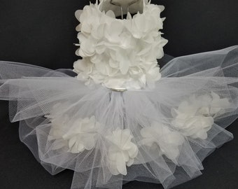 May include: White tulle pet dress with white fabric flowers. The dress has a layer of tulle and a layer of fabric flowers. The flowers are arranged in a circle around the waist.