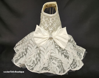 May include: A white lace dog dress with a satin bow and a rhinestone embellishment.
