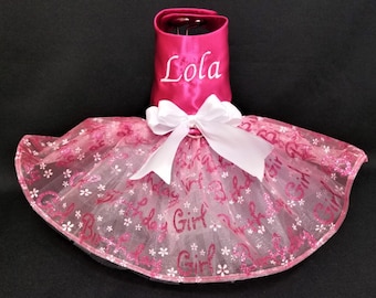 May include: Pink dog dress with a white tulle skirt and a pink satin bodice. The skirt has a pink glitter pattern with the words "Birthday Girl" repeated. The bodice has a white bow and the name "Lola" embroidered on the front.