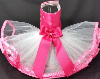 May include: A pink and white tulle tutu with a pink satin bow and polka dot trim. The tutu is designed for a pet.