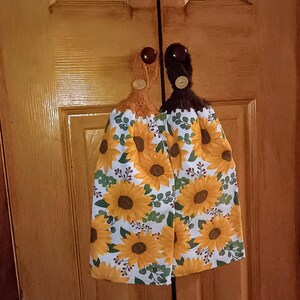 May include: Two crocheted dish towels with sunflower patterns and brown buttons. The towels are hanging on a wooden door.