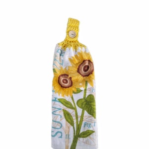 Puede incluir: Una toalla de cocina blanca con un dise&ntilde;o de girasol y el texto "Summer Sun" impreso en ella. La toalla tiene un lazo de crochet amarillo con un bot&oacute;n de madera.