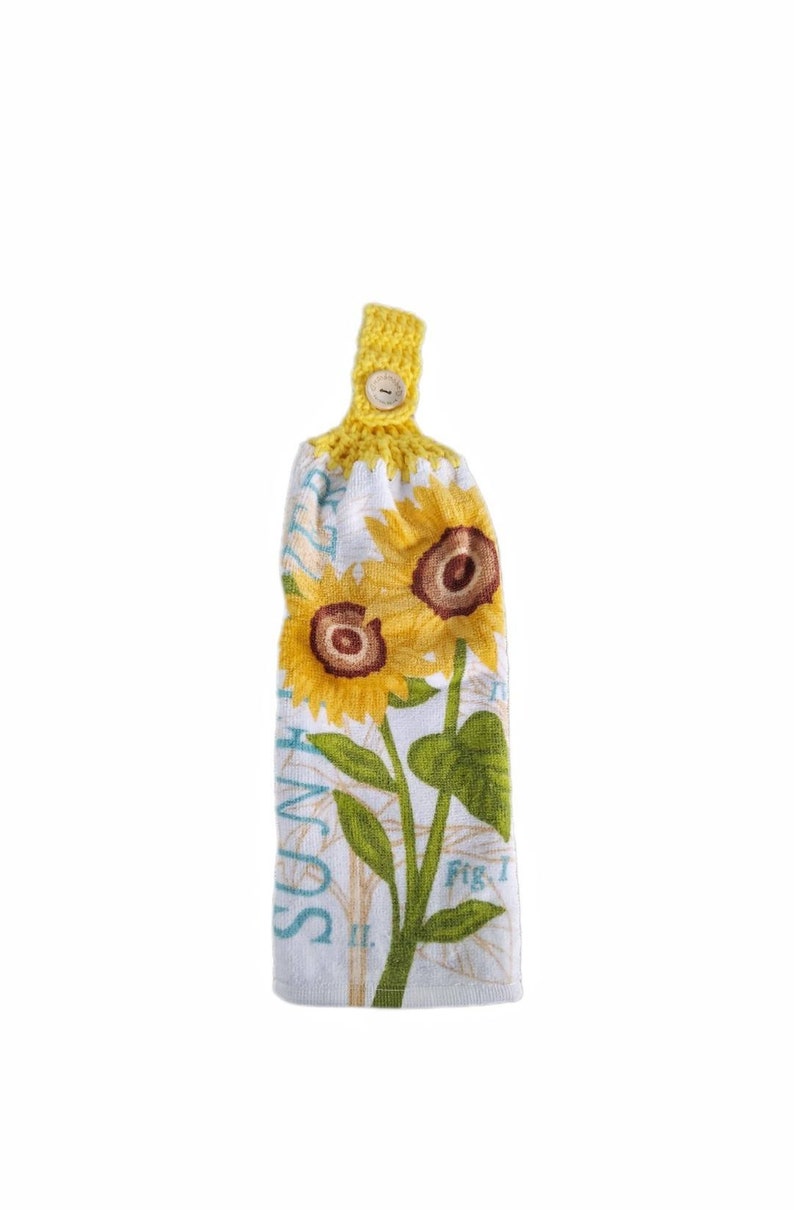 Puede incluir: Una toalla de cocina blanca con un dise&ntilde;o de girasol y el texto "Summer Sun" impreso en ella. La toalla tiene un lazo de crochet amarillo con un bot&oacute;n de madera.
