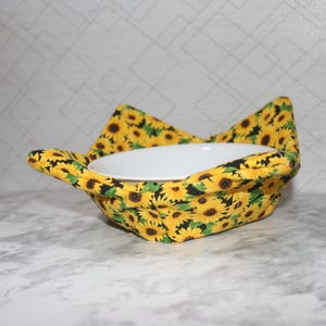 May include: A fabric bowl cozy featuring a vibrant sunflower pattern with yellow petals and dark brown centers on a black background. The quilted holder has four folded corners and cradles a plain white ceramic bowl in its center.