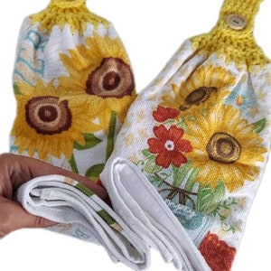 Puede incluir: Dos toallas de cocina blancas con estampado de girasoles y flores. Las toallas tienen la parte superior de crochet amarillo con un bot&oacute;n.
