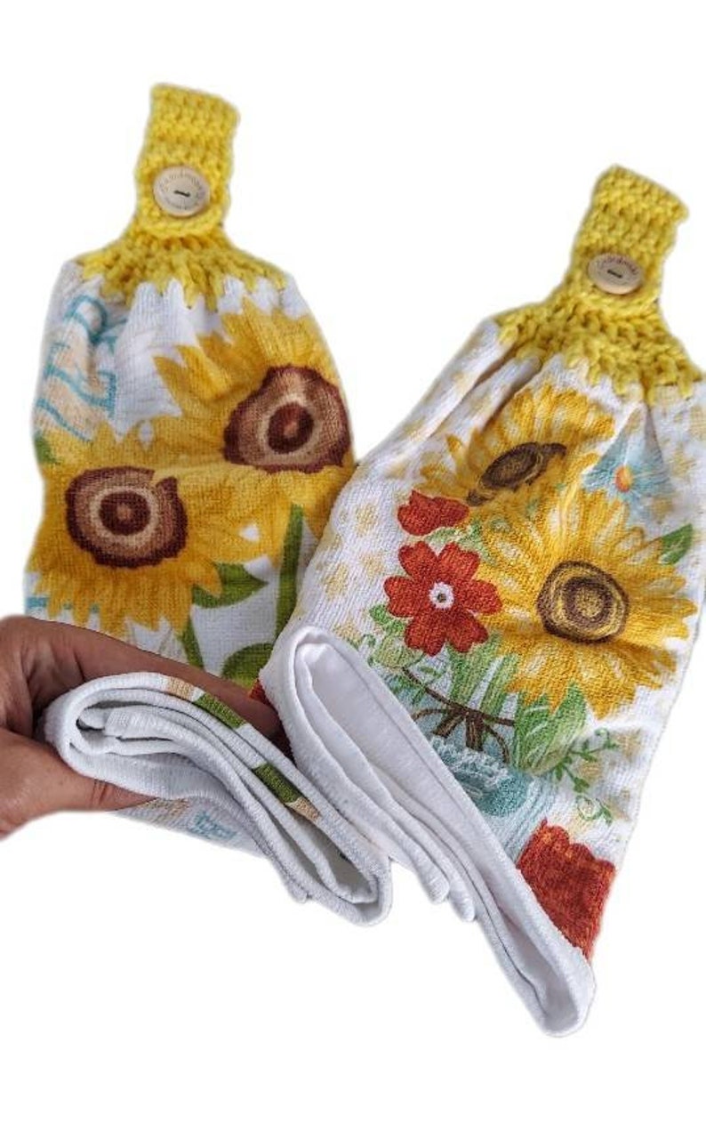 Puede incluir: Dos toallas de cocina blancas con estampado de girasoles y flores. Las toallas tienen la parte superior de crochet amarillo con un bot&oacute;n.