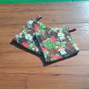 May include: Two square fabric pot holders featuring a festive pattern of bright red cardinals, green pine branches, and white birdhouses set against a dark brown wood grain background. Each quilted kitchen accessory includes a small fabric hanging loop.