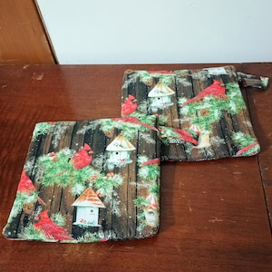 Puede incluir: Dos bolsas de tela con temática navideña. Las bolsas son marrones con un patrón de veta de madera y presentan cardenales rojos, ramas de pino y pequeñas casas para pájaros.