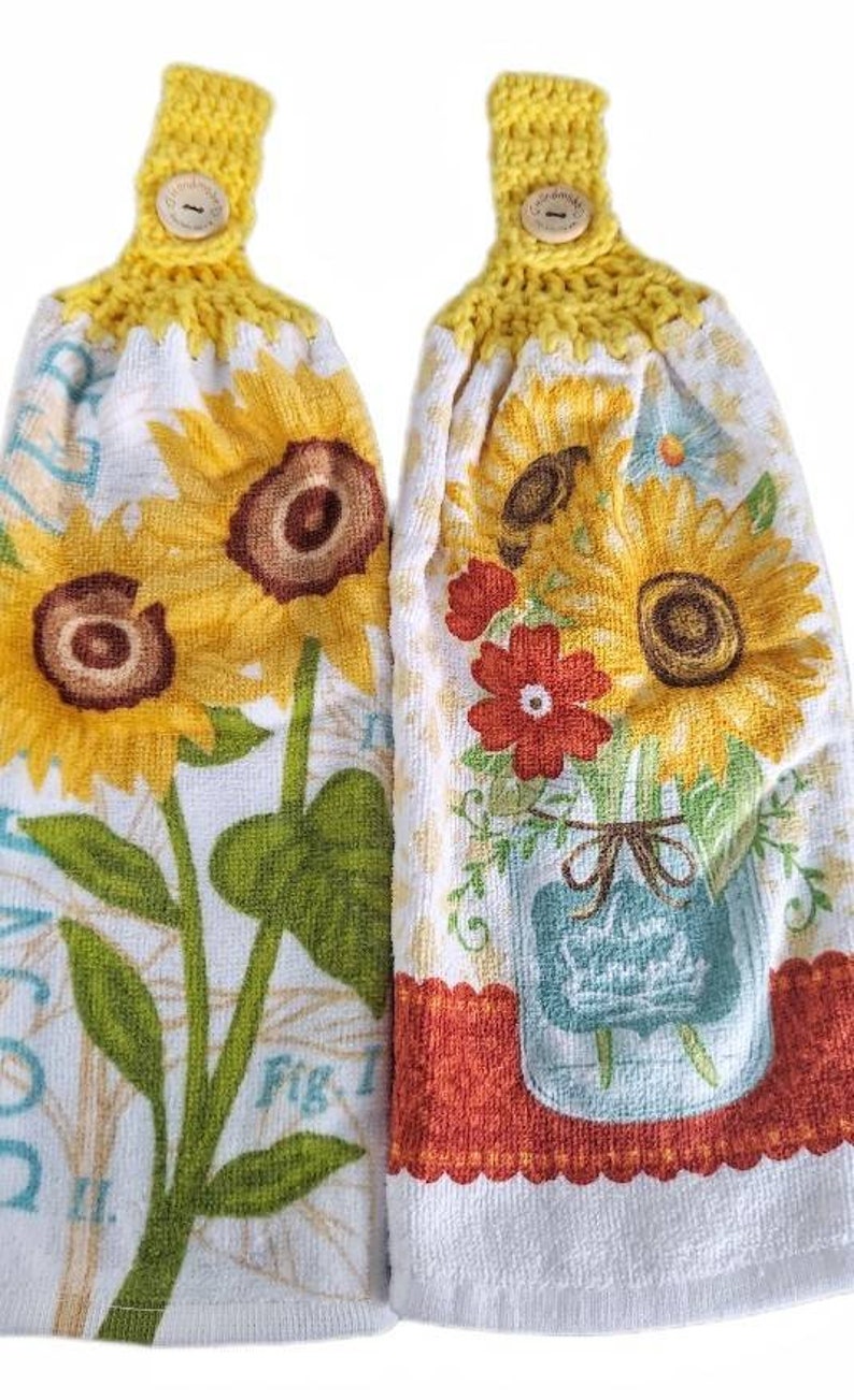 Puede incluir: Dos toallas de cocina de crochet amarillo con botones de madera. Una toalla presenta un dise&ntilde;o de girasol con el texto "Fig. 1", la otra presenta un dise&ntilde;o de girasol y flor roja con un tarro de cristal azul.
