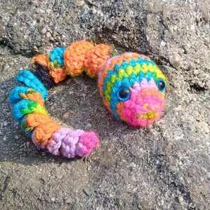 May include: A colorful, crocheted caterpillar toy with a rainbow of colors, including blue, orange, pink, and yellow. The caterpillar has large blue eyes and is posed on a textured, gray rock. The toy is handmade.