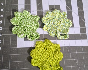 May include: Three crocheted four-leaf clover shapes in green, white, and yellow. The clovers are on a gray cutting mat.