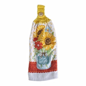 Puede incluir: Una toalla de cocina de crochet amarilla con un bot&oacute;n y un estampado de girasol. La toalla presenta un tarro de cristal lleno de girasoles y flores rojas.