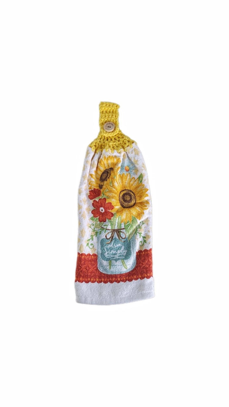 Puede incluir: Una toalla de cocina de crochet amarilla con un bot&oacute;n y un estampado de girasol. La toalla presenta un tarro de cristal lleno de girasoles y flores rojas.
