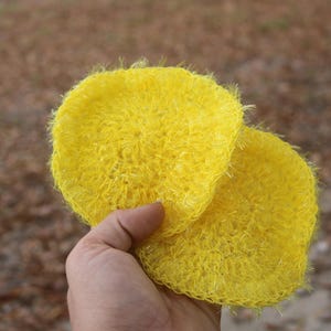 Puede incluir: Dos estropajos redondos de ganchillo de color amarillo brillante se sostienen en una mano. Los estropajos tienen un aspecto texturizado y en bucle. El fondo está borroso, con una mezcla de tonos marrones y verdes.
