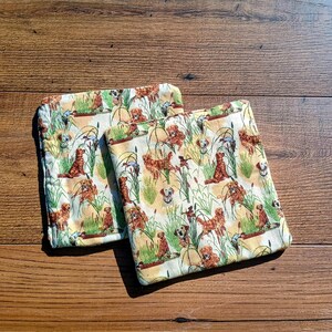 May include: Two square fabric coasters featuring a repeating pattern of golden retriever dogs sitting among green reeds and tall grasses on a light cream background. The dogs are depicted in various poses with brown fur and detailed textures.