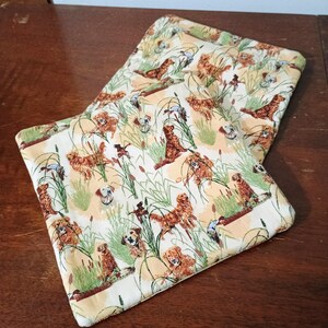 May include: Two square fabric pot holders featuring a repeating pattern of dogs, reeds, and ducks in brown, green, and cream. The pot holders are on a wooden surface.