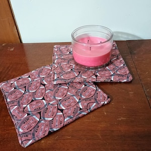 May include: Two square fabric coasters with a pattern of brown footballs with white stitching and the words "MVP" and "All Star" printed on them. A pink candle in a clear glass jar is sitting on top of the coasters.