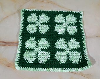 May include: A square crochet coaster featuring a dark forest green background with four light green and white variegated clover motifs arranged in a grid pattern, finished with a light green and white variegated border around the edges.