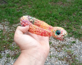Peut inclure: Un jouet serpent en crochet avec un œil vert et un corps multicolore, comprenant des nuances de rouge, d'orange, de jaune et de blanc.