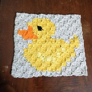 May include: A square crochet piece featuring a yellow duck design with an orange beak and a black eye, set against a light gray background. The item is crafted using a textured stitch pattern, creating a pixelated appearance for the duck motif.