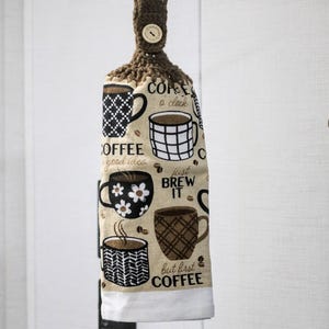 May include: A hanging kitchen towel with a brown crocheted top and button. The towel features various coffee cup designs and phrases like "Coffee o'clock" and "Just Brew It" on a beige background.
