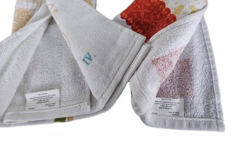 Puede incluir: Dos toallas de cocina blancas con estampados florales coloridos. Las toallas est&aacute;n dobladas por la mitad y muestran las instrucciones de cuidado en la etiqueta. La etiqueta dice 100% algod&oacute;n, lavar a m&aacute;quina con agua fr&iacute;a, secar en secadora a baja temperatura, ciclo delicado, no usar blanqueador, no planchar, hecho en China.